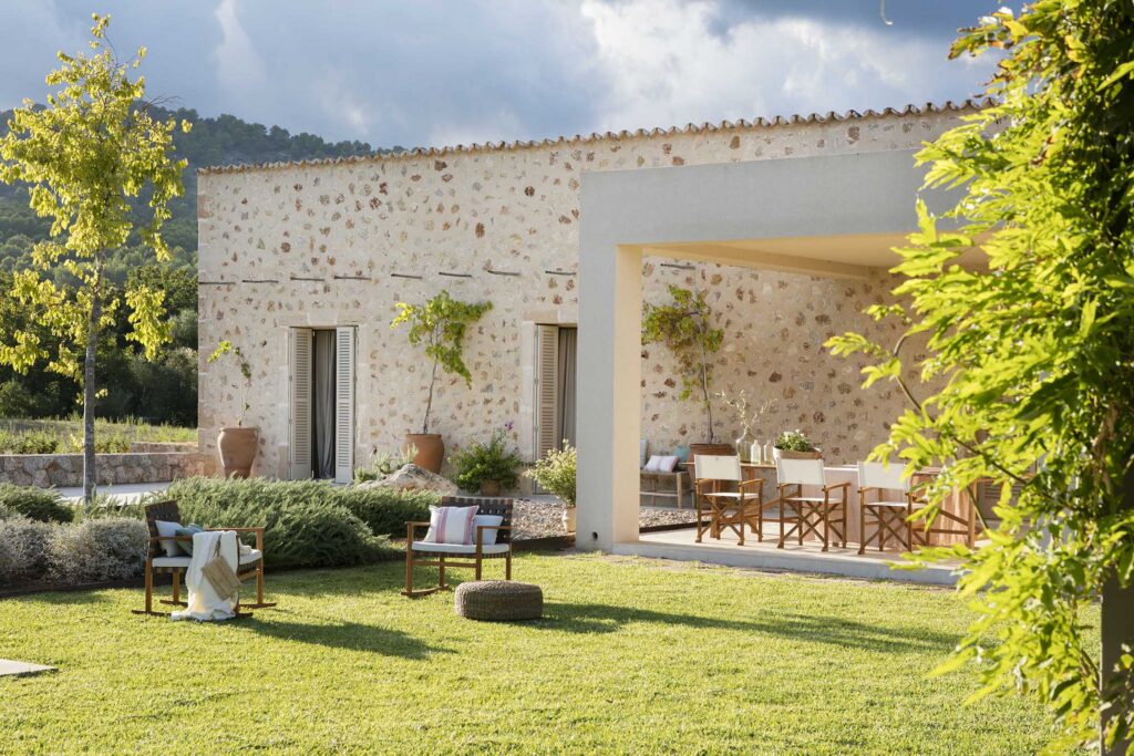 Fachada de piedra de Can Pelat con porche, sillas de madera y jardín de césped