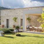 Fachada de piedra de Can Pelat con porche, sillas de madera y jardín de césped