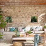Terraza cubierta con muro de piedra, sofá blanco corrido, cojines de colores y buganvilla