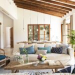 Salón con sofá blanco, cojines azules y mesa de centro de madera, con la cocina al fondo