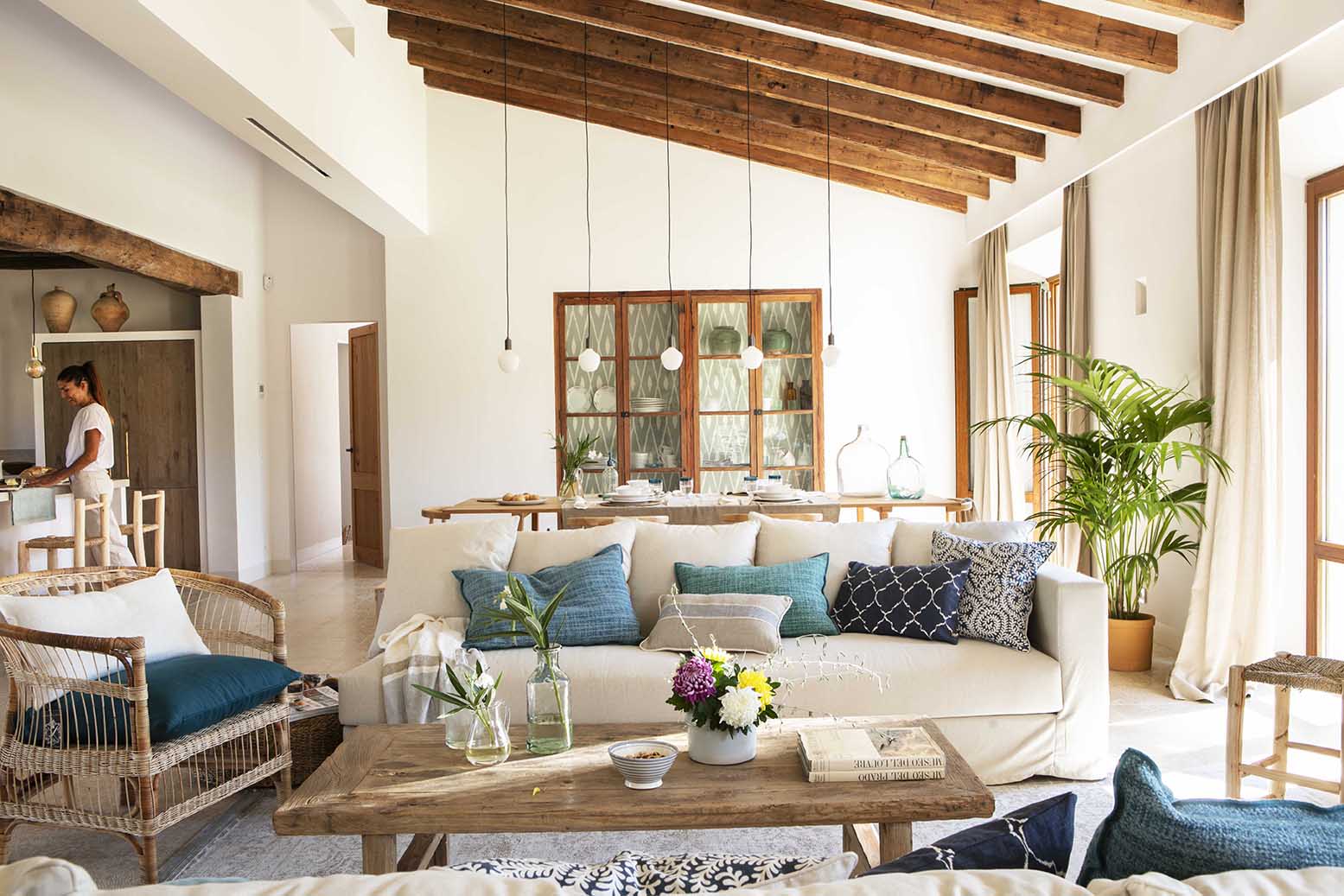 Salón con sofá blanco, cojines azules y mesa de centro de madera, con la cocina al fondo