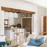 Salón abierto a la cocina con gran viga de madera, barra blanca, taburetes y armario de madera