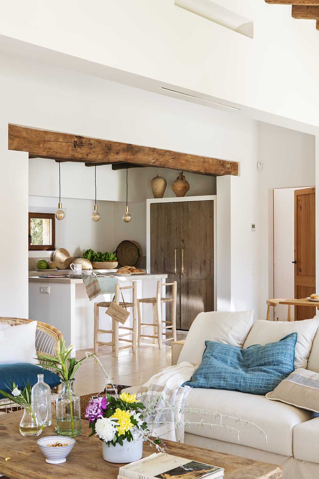 Salón abierto a la cocina con gran viga de madera, barra blanca, taburetes y armario de madera