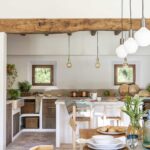 Cocina rústica con gran viga de madera, ventanas pequeñas, isla blanca y mesa puesta en primer plano