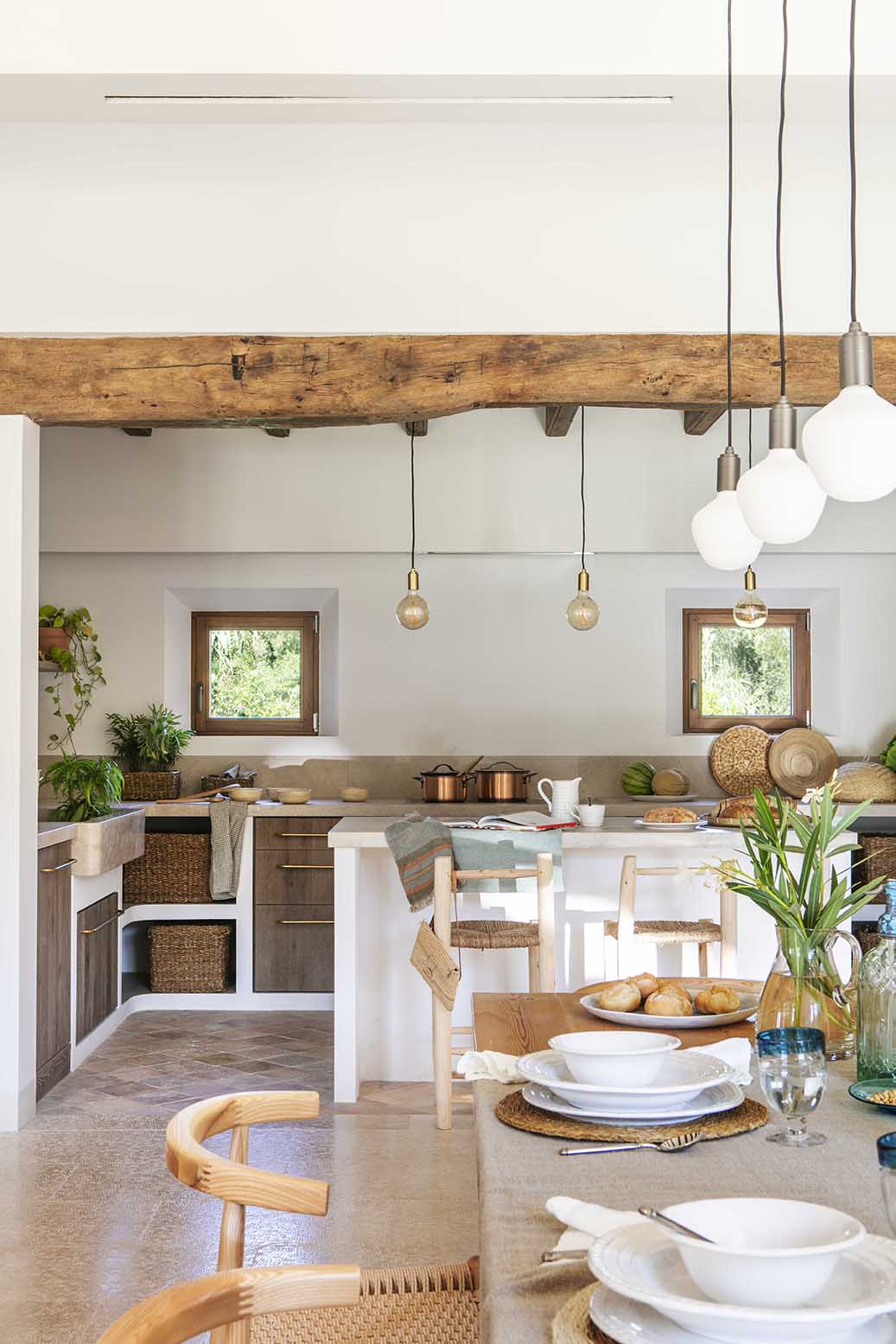 Cocina rústica con gran viga de madera, ventanas pequeñas, isla blanca y mesa puesta en primer plano