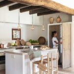 Cocina con isla blanca, encimera de piedra, taburetes de madera, frigorífico integrado y baldosas de barro
