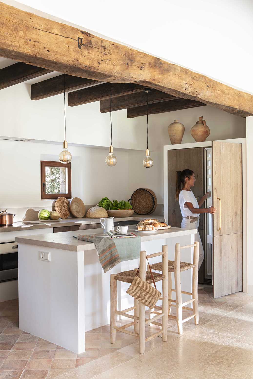 Cocina con isla blanca, encimera de piedra, taburetes de madera, frigorífico integrado y baldosas de barro