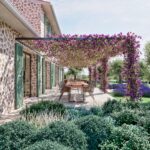 Esstisch im Freien unter einer Pergola mit violetten Blüten neben einer Steinfassade mit grünen Fensterläden