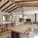 Offene Küche mit heller Kochinsel, Barhockern aus Holz, Esstisch, Hängekamin und sichtbaren Holzbalken