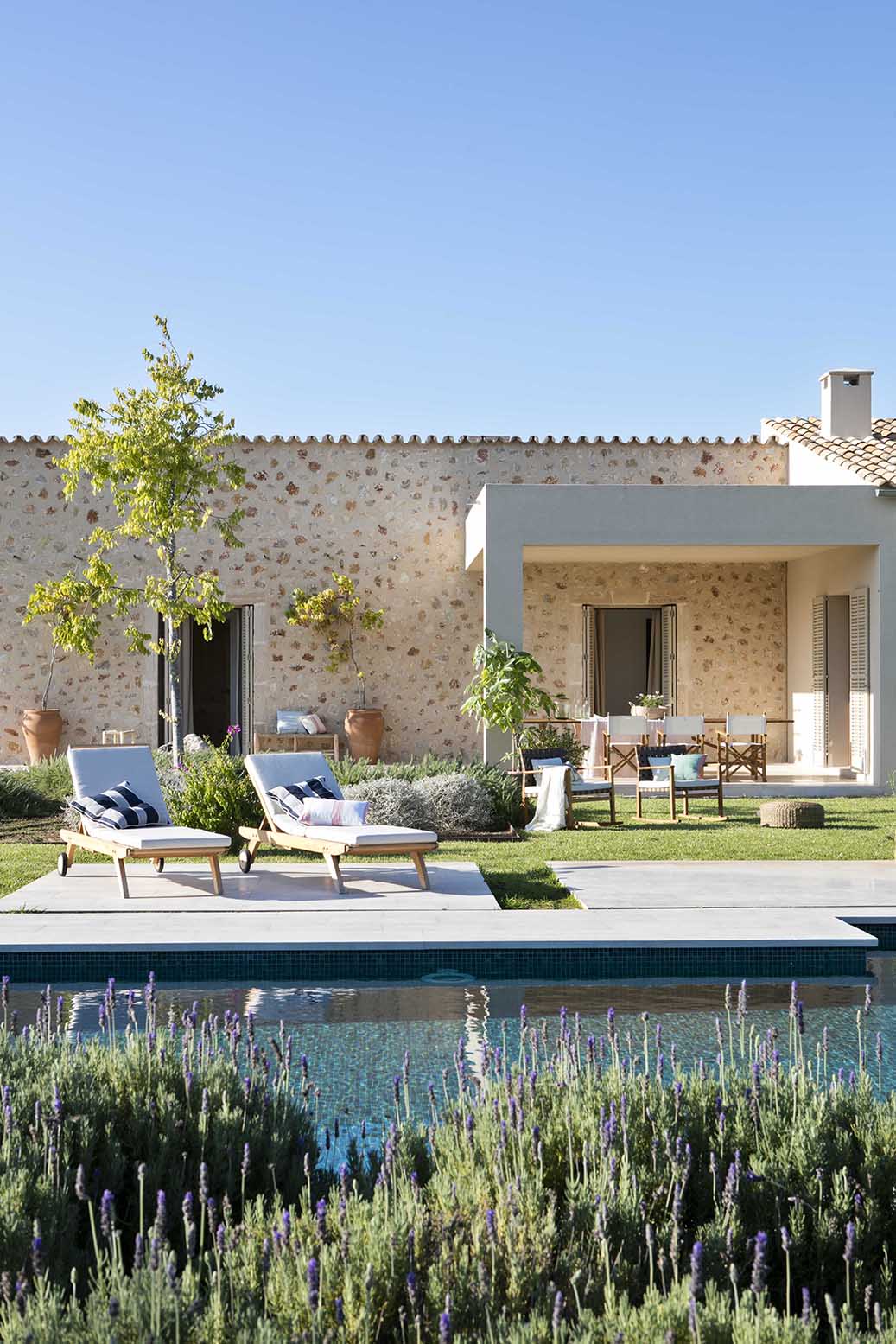 Fachada de piedra vista desde la piscina, con tumbonas, lavanda en primer plano y cielo azul despejado
