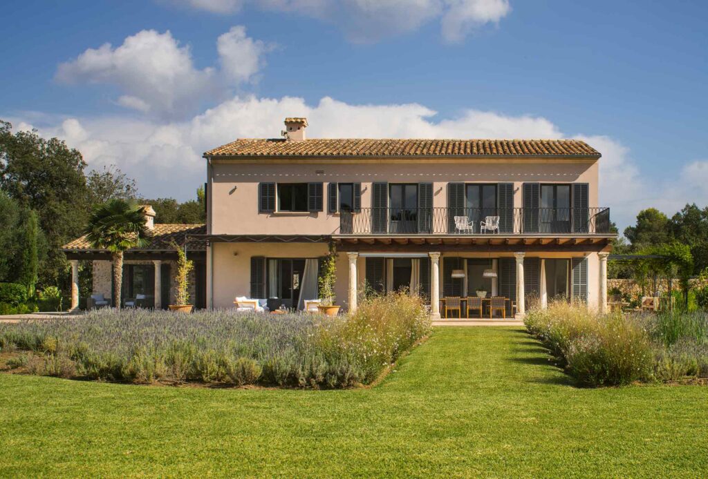 Fachada al jardín con balcón de forja, porche de columnas de piedra y césped con lavanda