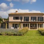 Fachada al jardín con balcón de forja, porche de columnas de piedra y césped con lavanda