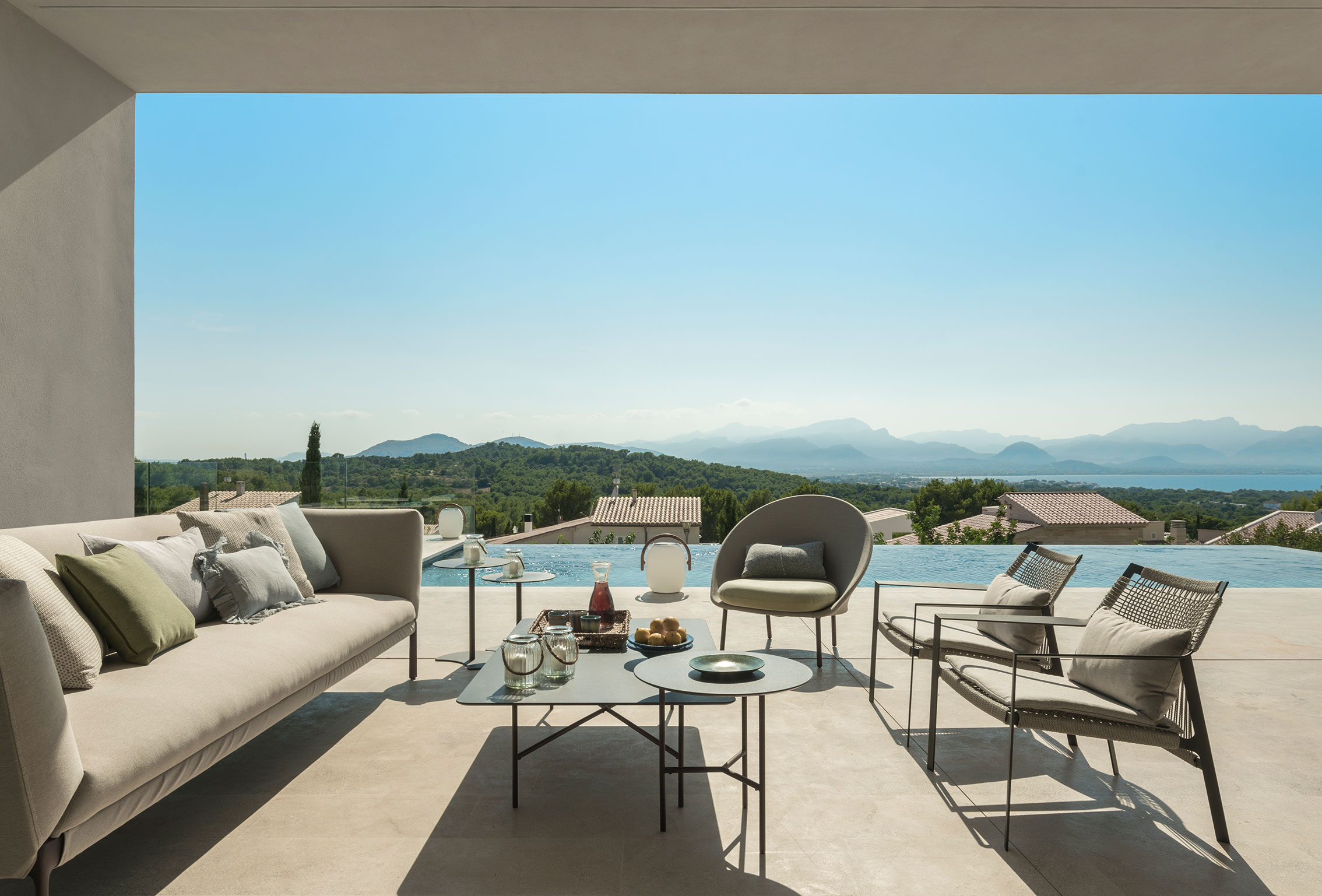 Beige sofa and braided rope armchairs under the porch, with an infinity pool and mountains behind