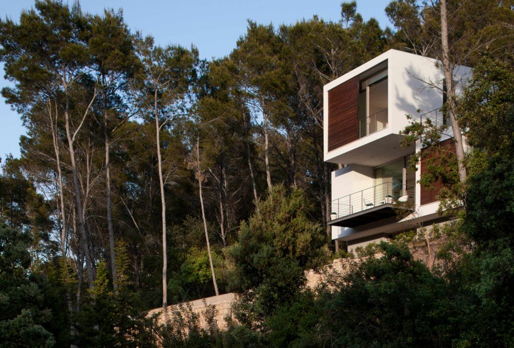 Vista de la casa desde el bosque, con volúmenes blancos y madera asomando entre pinos y vegetación