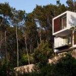 Vista de la casa desde el bosque, con volúmenes blancos y madera asomando entre pinos y vegetación