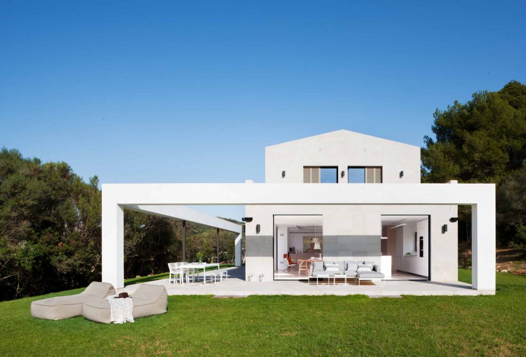 Fachada blanca con pórtico, salón y cocina abiertos al jardín, puffs de exterior y comedor bajo porche
