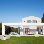 Fachada blanca con pórtico, salón y cocina abiertos al jardín, puffs de exterior y comedor bajo porche