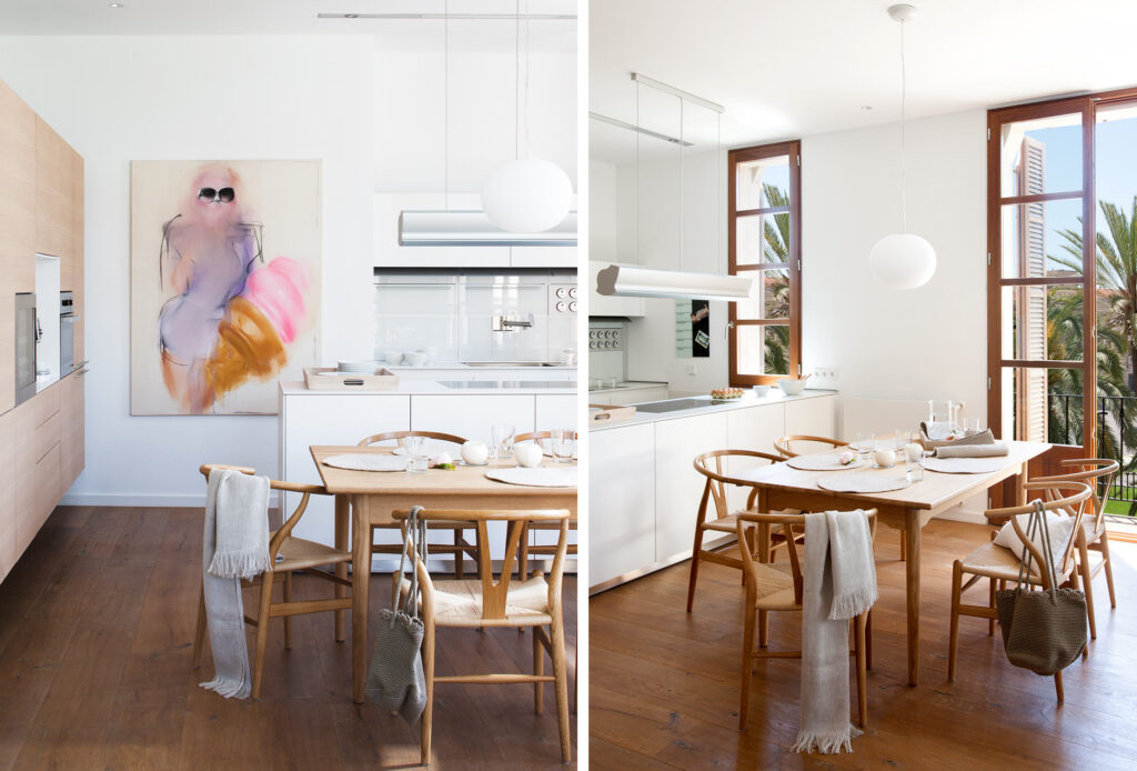 Cocina blanca con comedor, sillas y mesa de madera, armarios de madera clara y ventana con palmeras