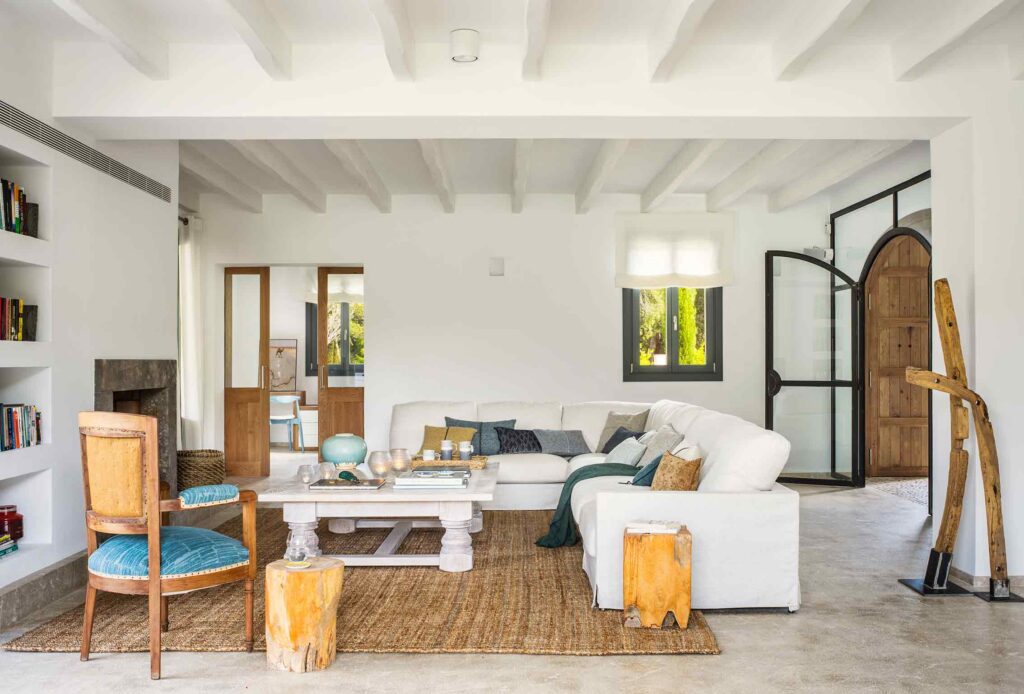 Salón con vigas blancas, sofá blanco, suelo continuo claro y puerta de hierro y cristal en arco