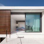 Terrasse mit hellem Boden vor einem Schlafzimmer mit großem Fenster, türkisfarbenem Vorhang und Holzgitter