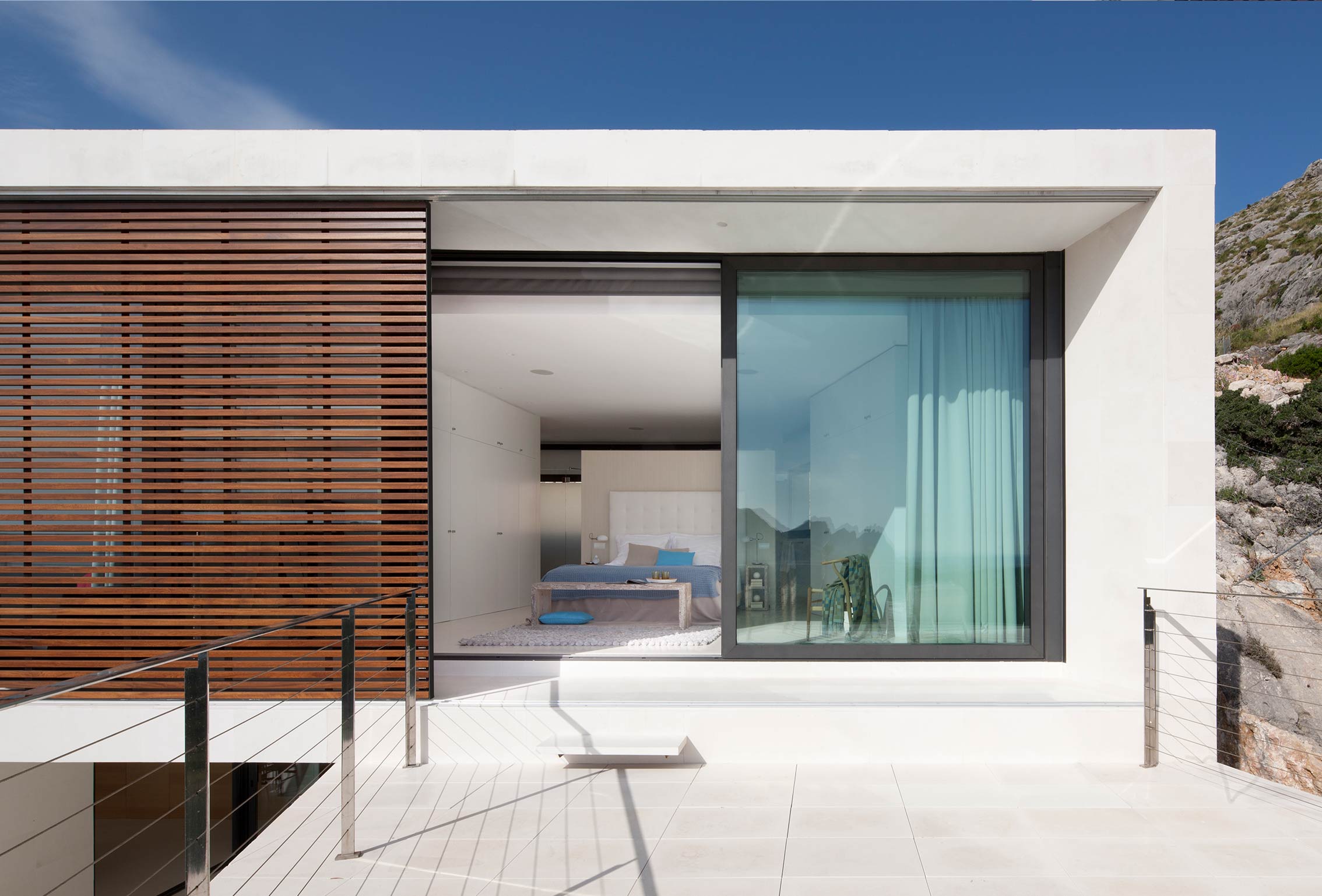 Terrasse mit hellem Boden vor einem Schlafzimmer mit großem Fenster, türkisfarbenem Vorhang und Holzgitter