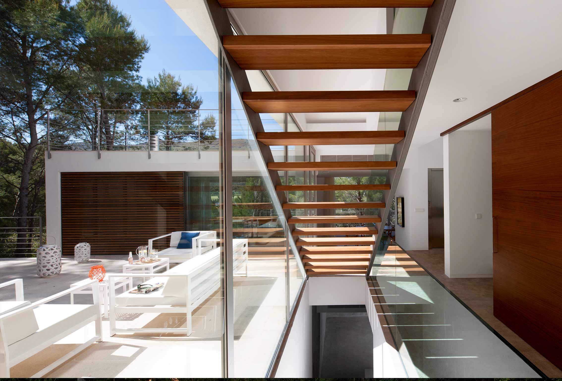 Escalera de peldaños de madera junto a cristalera con vistas a la terraza de sofás blancos