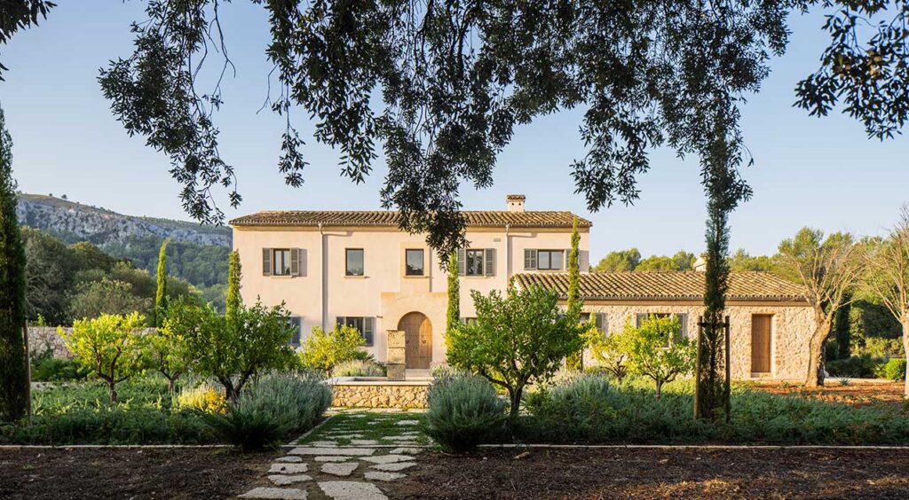 Fachada de Can Bosc, casa de campo de piedra y revoco claro con tejas y jardín mediterráneo