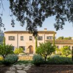 Fachada de Can Bosc, casa de campo de piedra y revoco claro con tejas y jardín mediterráneo