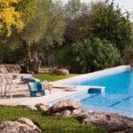 Piscina azul con borde de piedra clara, tumbonas blancas, rocas y arbolado del entorno al fondo