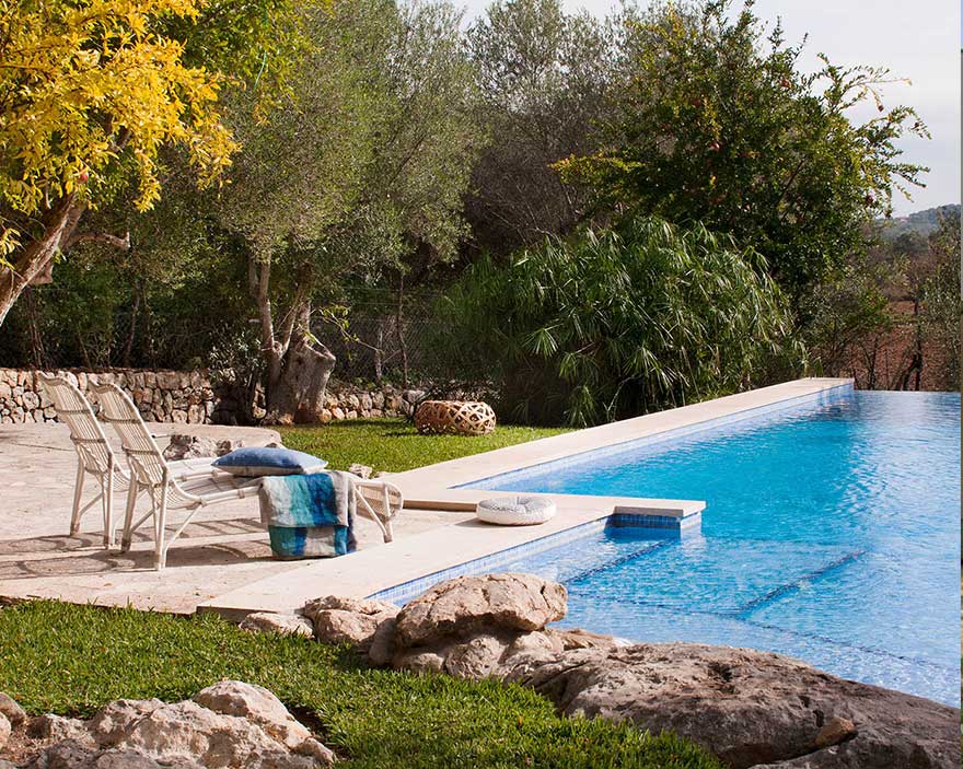 Piscina azul con borde de piedra clara, tumbonas blancas, rocas y arbolado del entorno al fondo