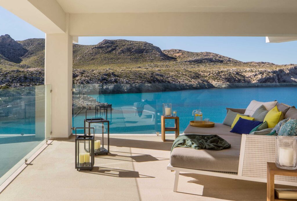 Terraza cubierta con sofá lounge, cojines de colores, linternas y barandilla de cristal frente a la cala