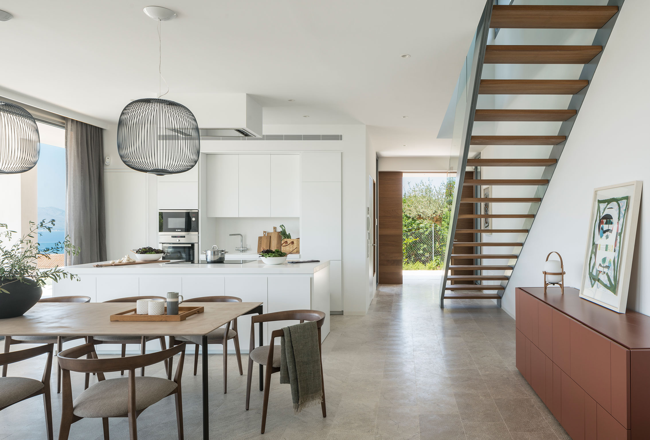 White kitchen with island open to the dining area, timber staircase and light stone floor