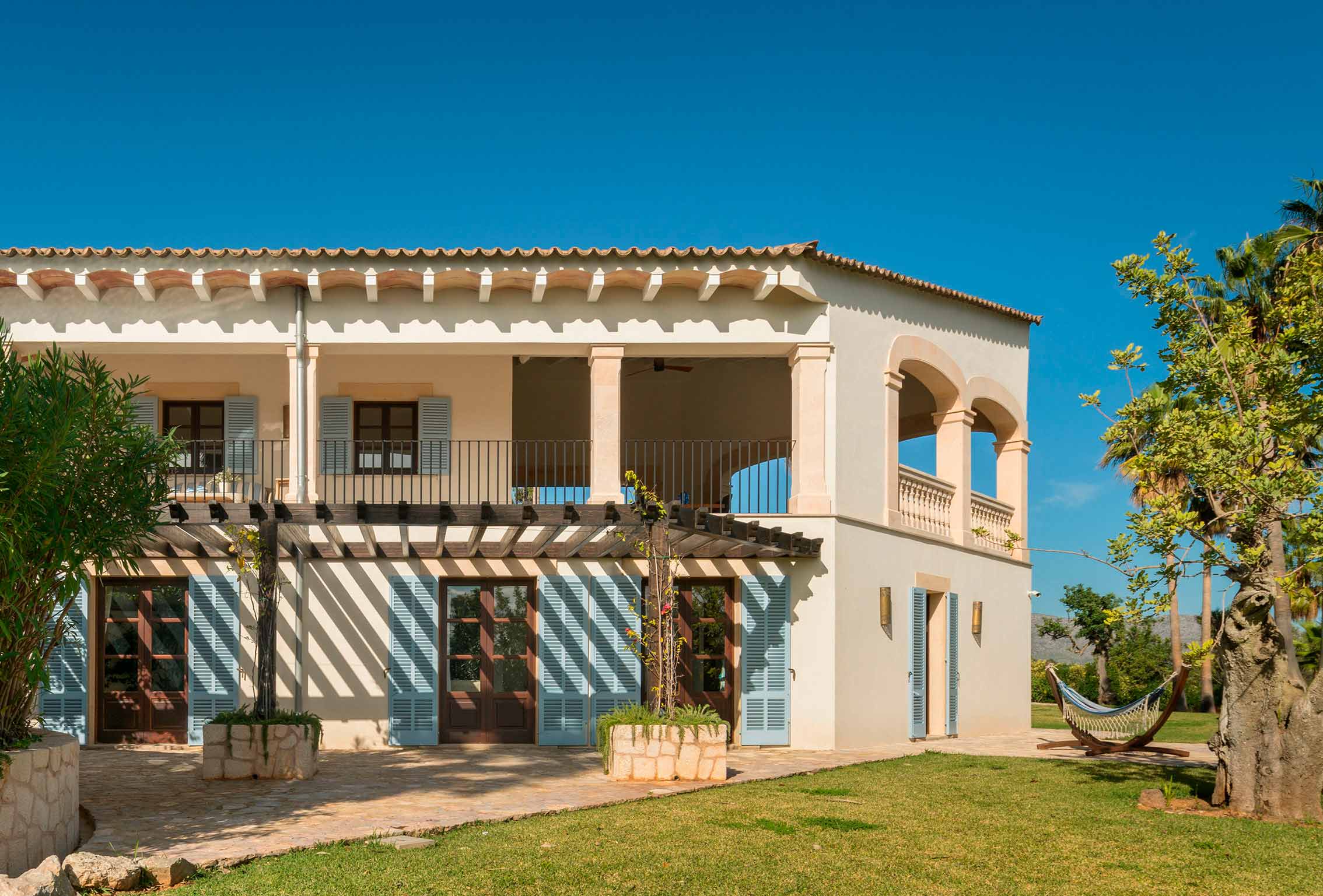 Fachada de dos plantas con terraza cubierta, persianas azules, carpintería de madera y pérgola sobre patio empedrado