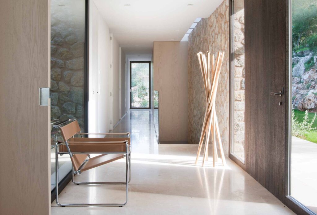 Pasillo con muro de piedra, suelo claro pulido, silla de cuero y perchero de madera