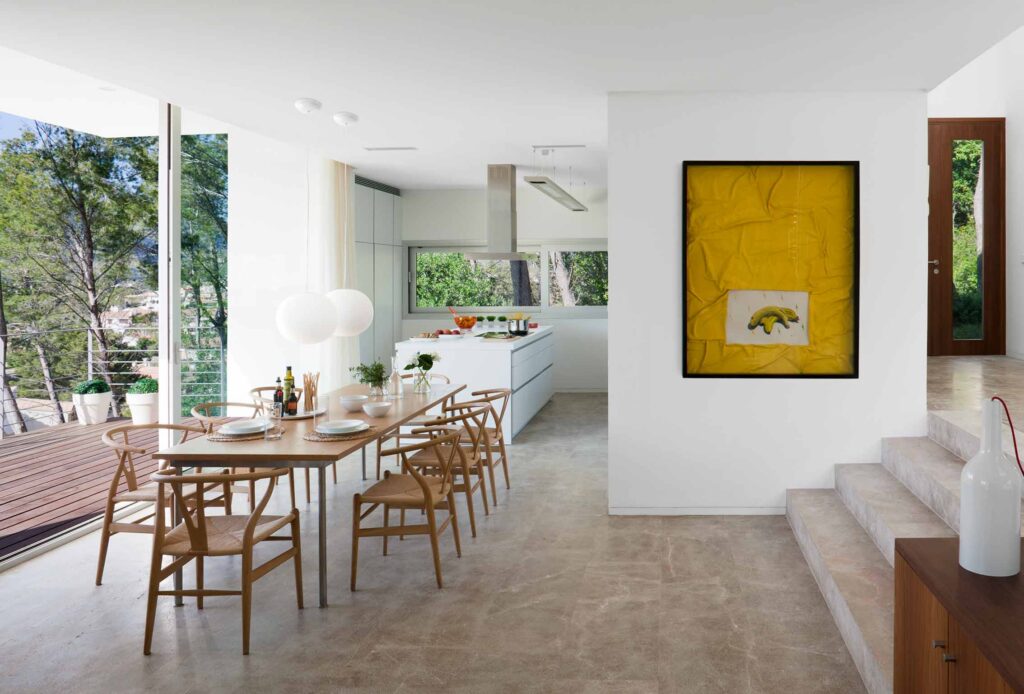 Comedor con mesa y sillas de madera, cocina blanca al fondo, cuadro amarillo y escalones de piedra