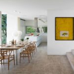 Comedor con mesa y sillas de madera, cocina blanca al fondo, cuadro amarillo y escalones de piedra