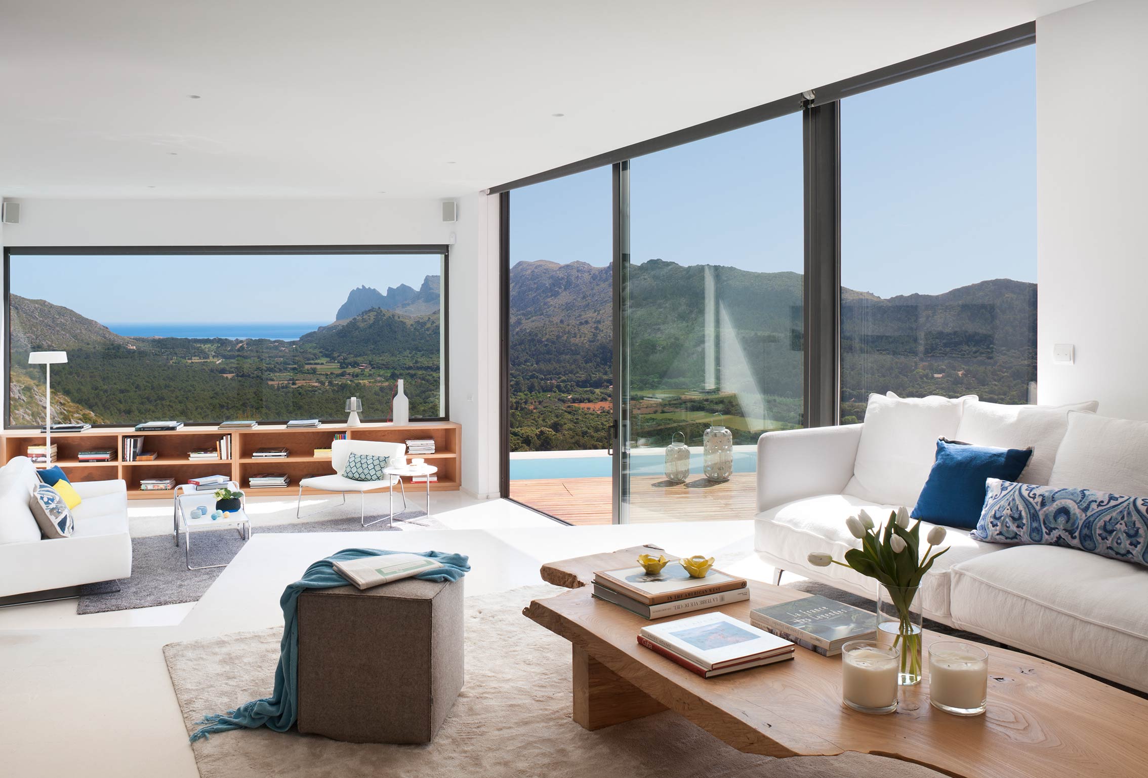 Wohnzimmer mit weißen Sofas, Holztisch und großen Fenstern mit Blick auf Berge und Meer