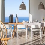 Comedor con mesa de madera, sillas blancas, lámparas colgantes grises y el mar azul al fondo