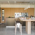 Cocina abierta con armarios de roble claro, isla con barra de madera, taburetes altos y vinoteca integrada
