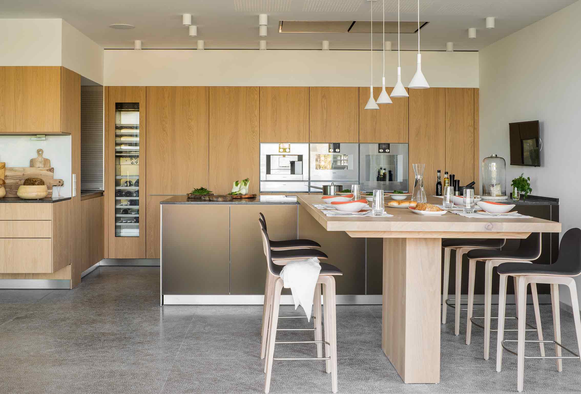 Cocina abierta con armarios de roble claro, isla con barra de madera, taburetes altos y vinoteca integrada