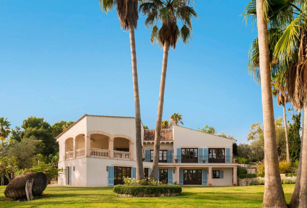 Casa de dos plantas entre altas palmeras y césped, con terraza de arcos y persianas azules