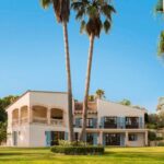 Casa de dos plantas entre altas palmeras y césped, con terraza de arcos y persianas azules