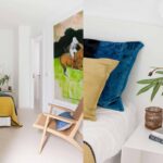 Schlafzimmer mit senffarbener Tagesdecke, Holzsessel und großem Bild, dazu Detail blauer und senffarbener Kissen