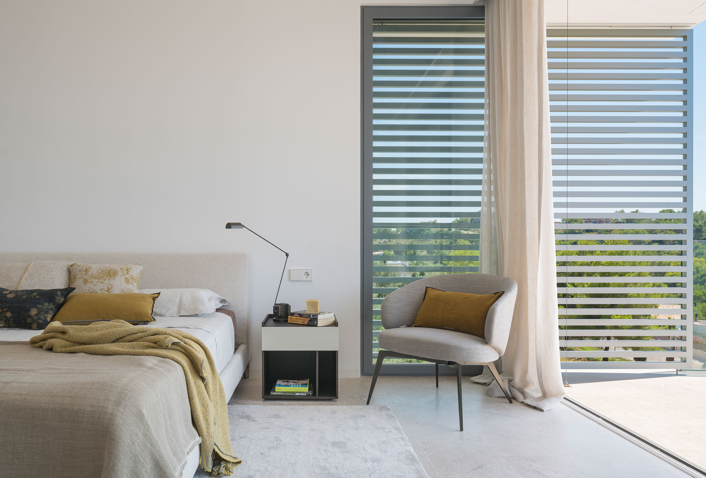 Bedroom with mustard cushions, grey armchair, slatted screen and light curtain beside the terrace