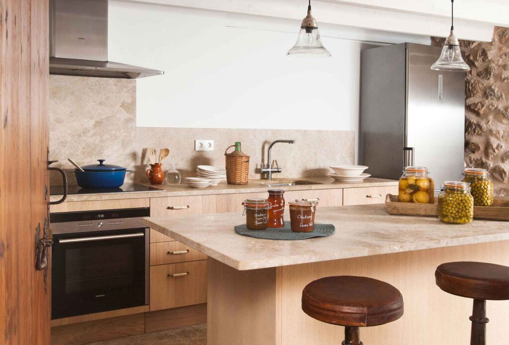 Cocina con muebles de madera clara, encimera y salpicadero de piedra beige, isla con taburetes de cuero
