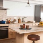 Cocina con muebles de madera clara, encimera y salpicadero de piedra beige, isla con taburetes de cuero