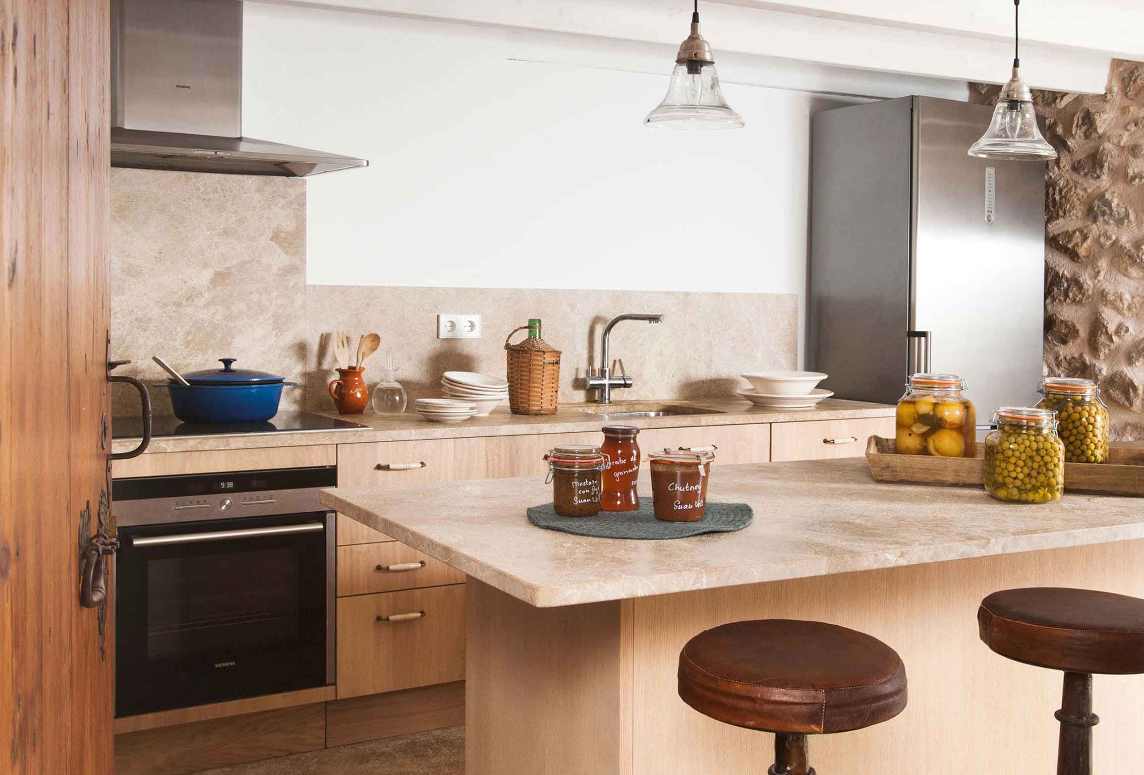Cocina con muebles de madera clara, encimera y salpicadero de piedra beige, isla con taburetes de cuero