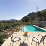 Langgestreckter türkisfarbener Pool neben einer Steinmauer, mit Terrasse, zwei Stühlen und Bergen dahinter