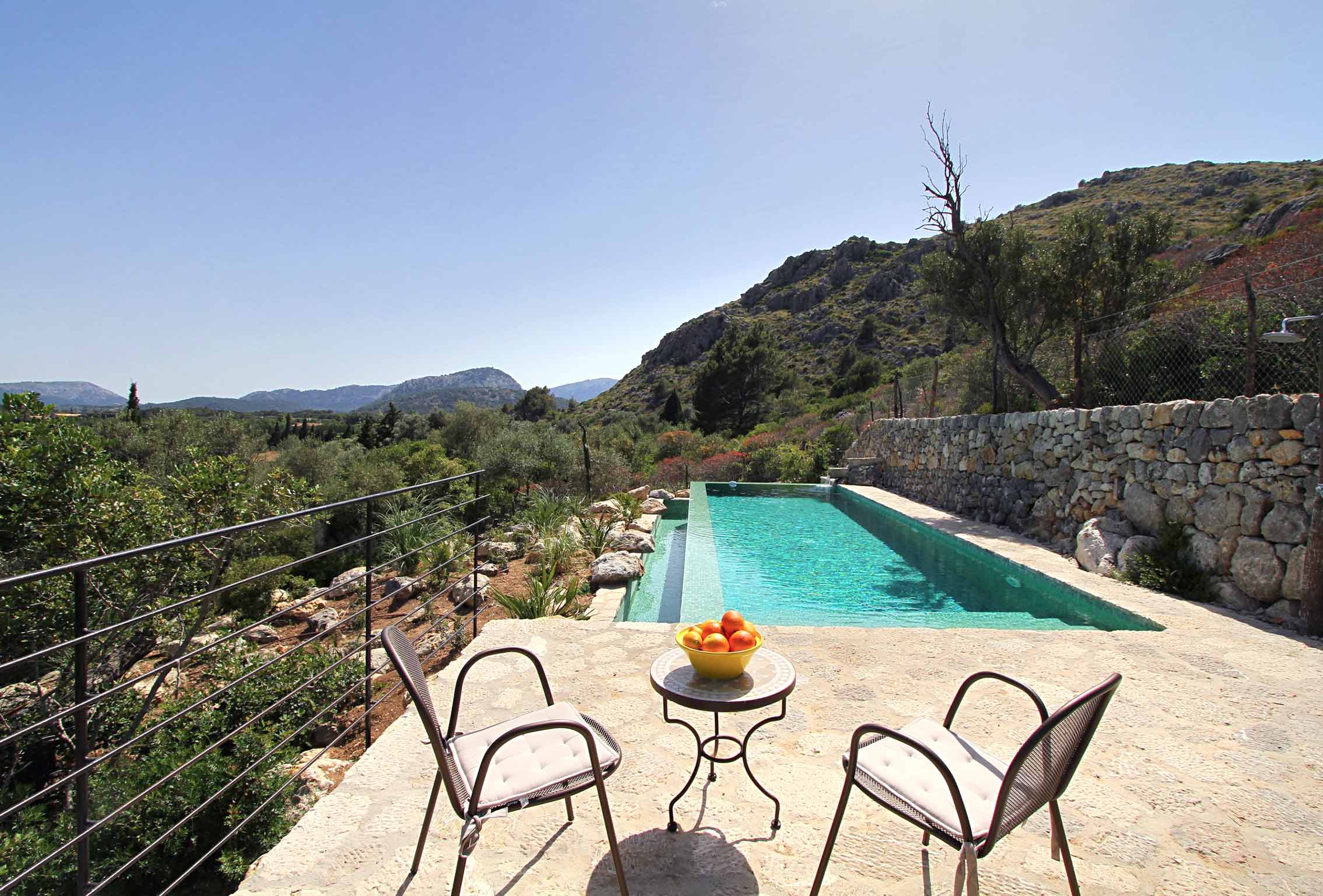 Langgestreckter türkisfarbener Pool neben einer Steinmauer, mit Terrasse, zwei Stühlen und Bergen dahinter