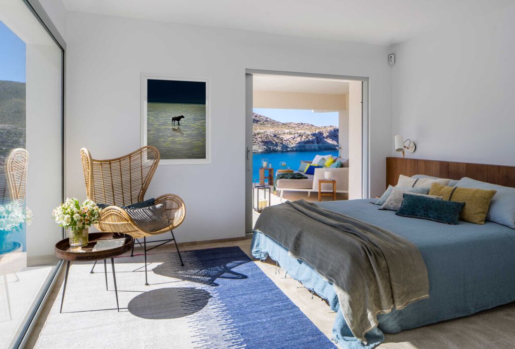 Dormitorio con cabecero de madera, ropa de cama azul, butaca de ratán y salida a la terraza